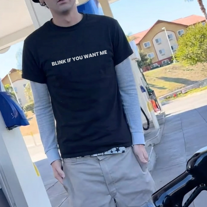 A man wearing a black graphic t-shirt with the text 'BLINK IF YOU WANT ME' stands at a gas station pump. He is also wearing a gray beanie, a light gray long-sleeve shirt underneath, khaki shorts, white socks, and slip-on shoes.