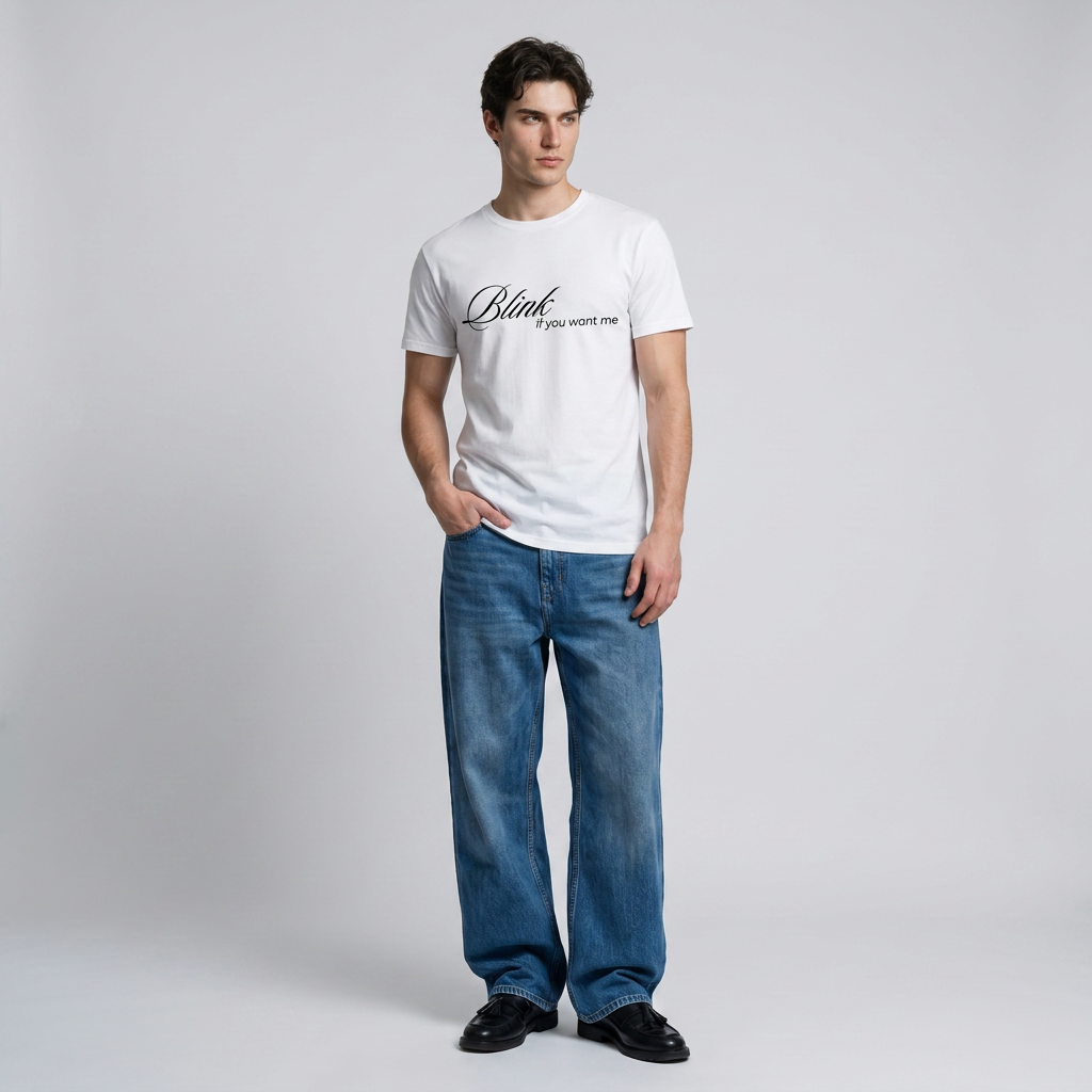 A man modeling a white crew-neck t-shirt with black script text on the chest, paired with blue jeans and black shoes, standing against a light gray studio background.