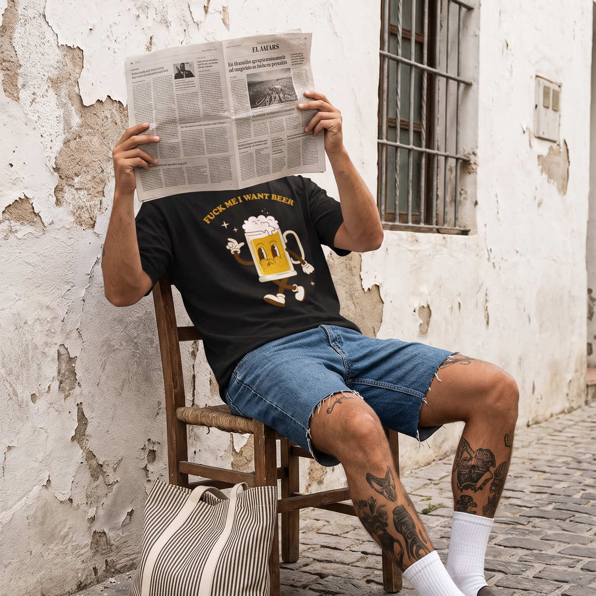 A man sitting on a wooden chair against a weathered white wall, holding a newspaper in front of his face. He is wearing a black graphic t-shirt, blue denim shorts, white socks, and brown leather loafers, with a striped tote bag resting on the floor beside him.